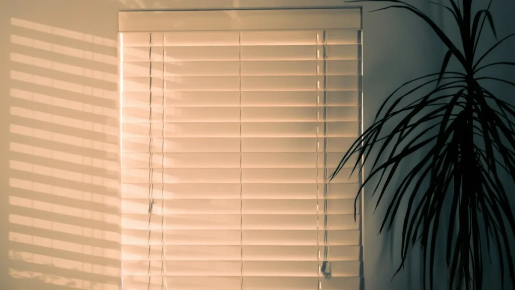Persiana horizontal branca fechada em janela de sala iluminada, ao lado de uma planta